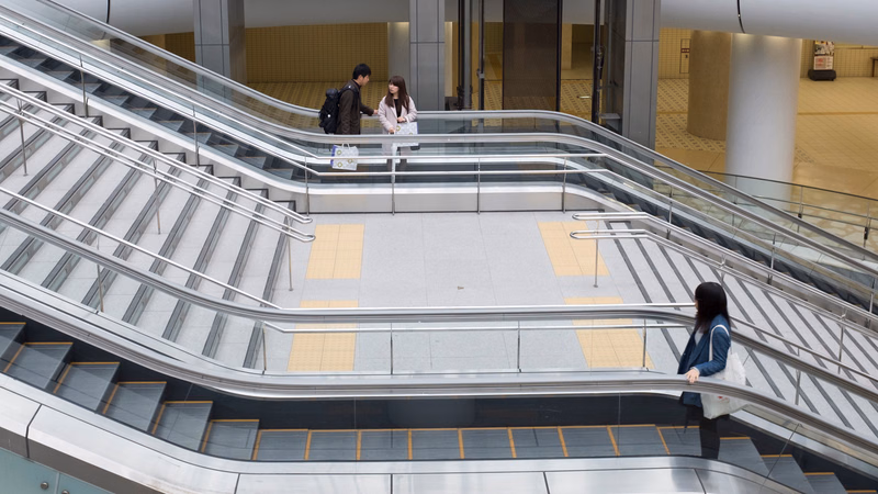 Two people walk on an escalator in a bright, modern indoor shopping mall setting.