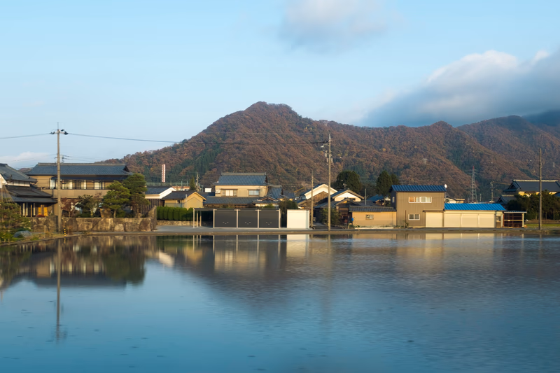 A serene village scene with a lake in the foreground and mountains in the background.