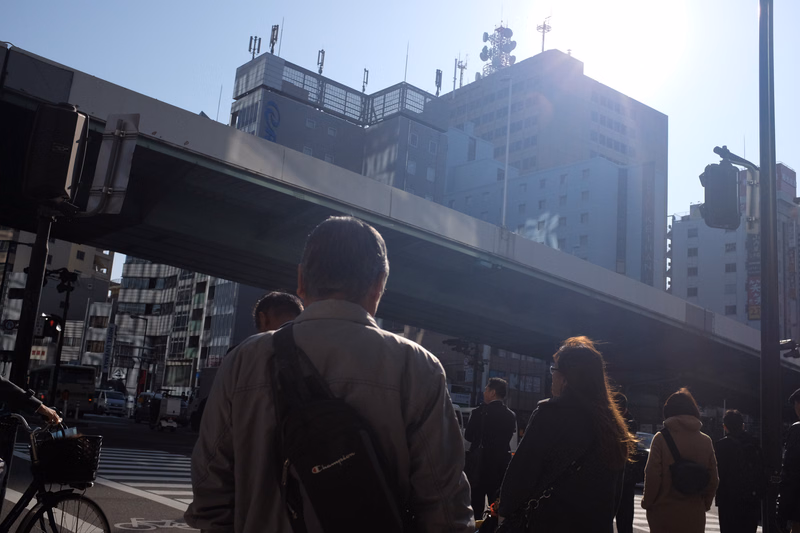 Several pedestrians walk under a large elevated overpass in a busy urban setting with tall buildings in the background.