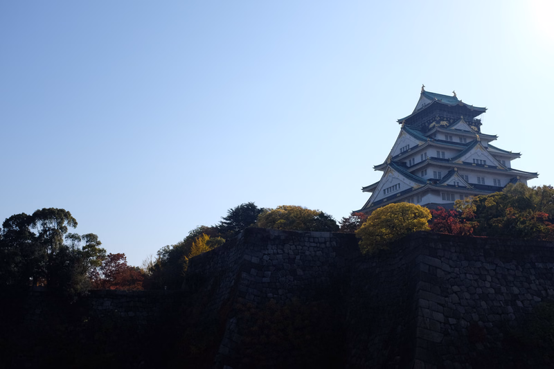 A traditional Japanese castle with tiered roofs is visible above a dark stone wall against a clear blue sky.