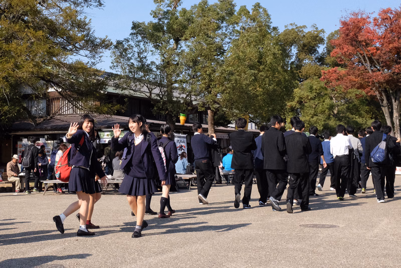 A group of students in school uniforms walks across a paved area surrounded by lush green and red trees on a sunny day.