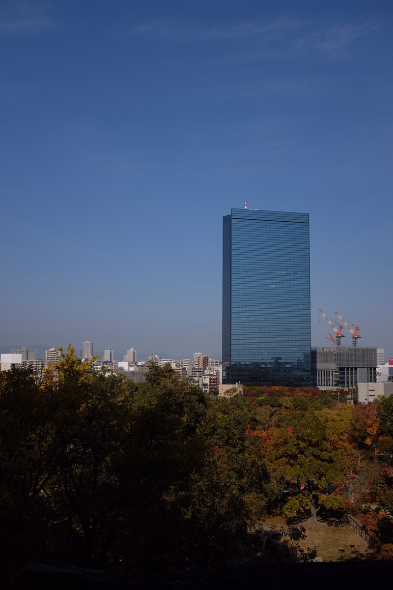 A tall blue glass skyscraper stands against a clear blue sky, surrounded by trees and other city buildings.