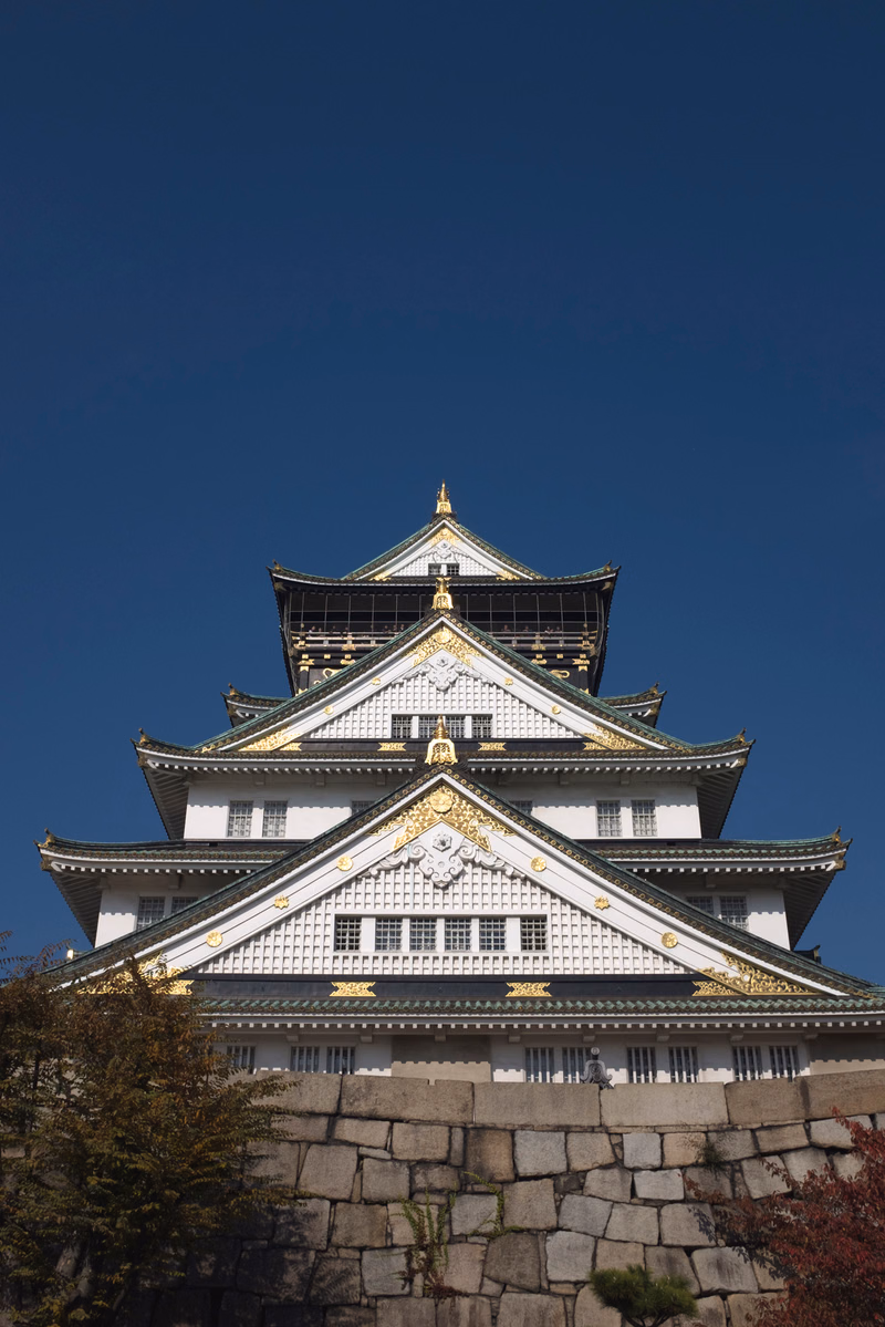 A large, traditional Japanese castle with white walls and dark tiled roofs stands behind a stone wall against a clear blue sky.