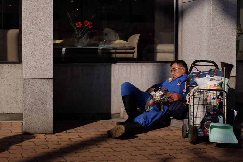 An elderly man in a blue uniform sits in a wheelchair outside a modern building entrance.