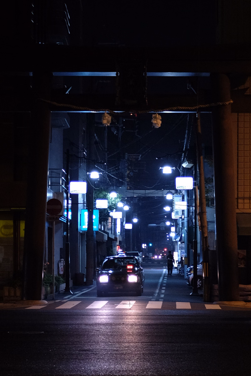 A nighttime view down a city street with illuminated traffic lights, street lamps, and a car driving through a crosswalk.