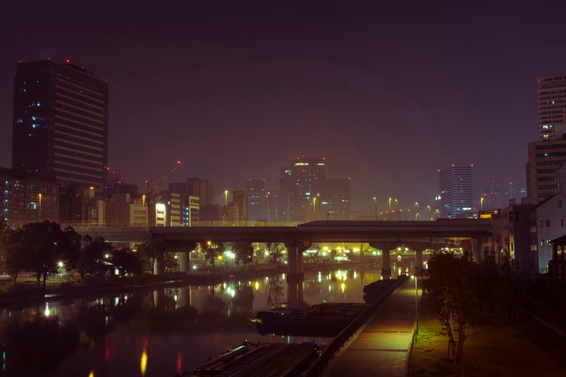 A nighttime cityscape view featuring a bridge crossing over a river with illuminated buildings in the background.