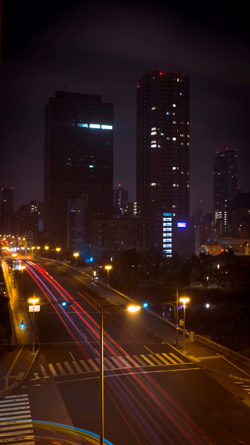 A nighttime cityscape view showing tall modern buildings illuminated against the dark sky, with streaks of car lights on the street below.