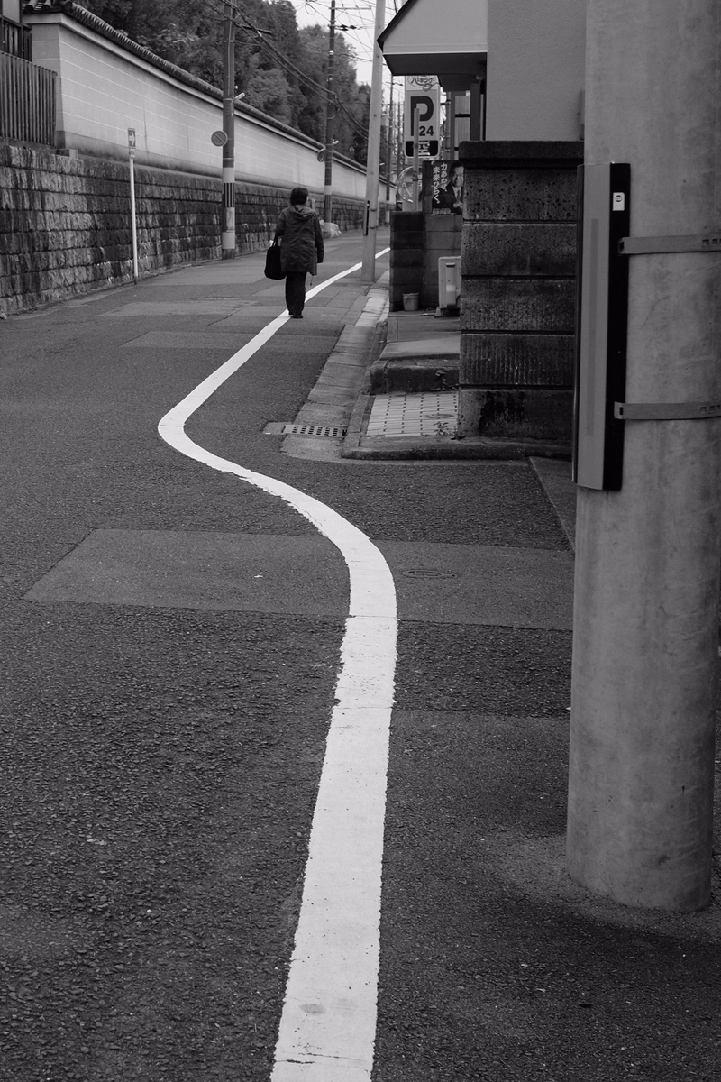 A person walks along a sidewalk next to a curb, with a white line marking the edge of the pavement.