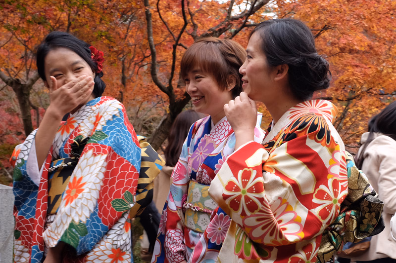 Three women wearing colorful kimono stand outdoors among trees with autumn foliage.