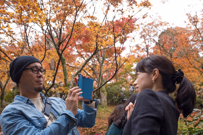 A man and a woman stand outdoors in an autumn park, with the man holding up a blue smartphone.