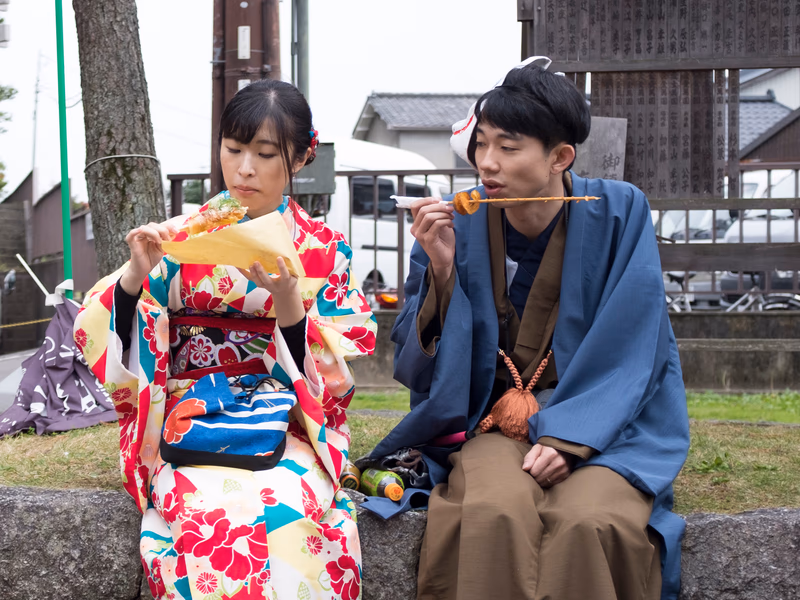 A woman in a kimono and a man in a kimono are sitting on a stone ledge, talking and eating from a stick. The image is set outdoors near a wooden structure and a tree.