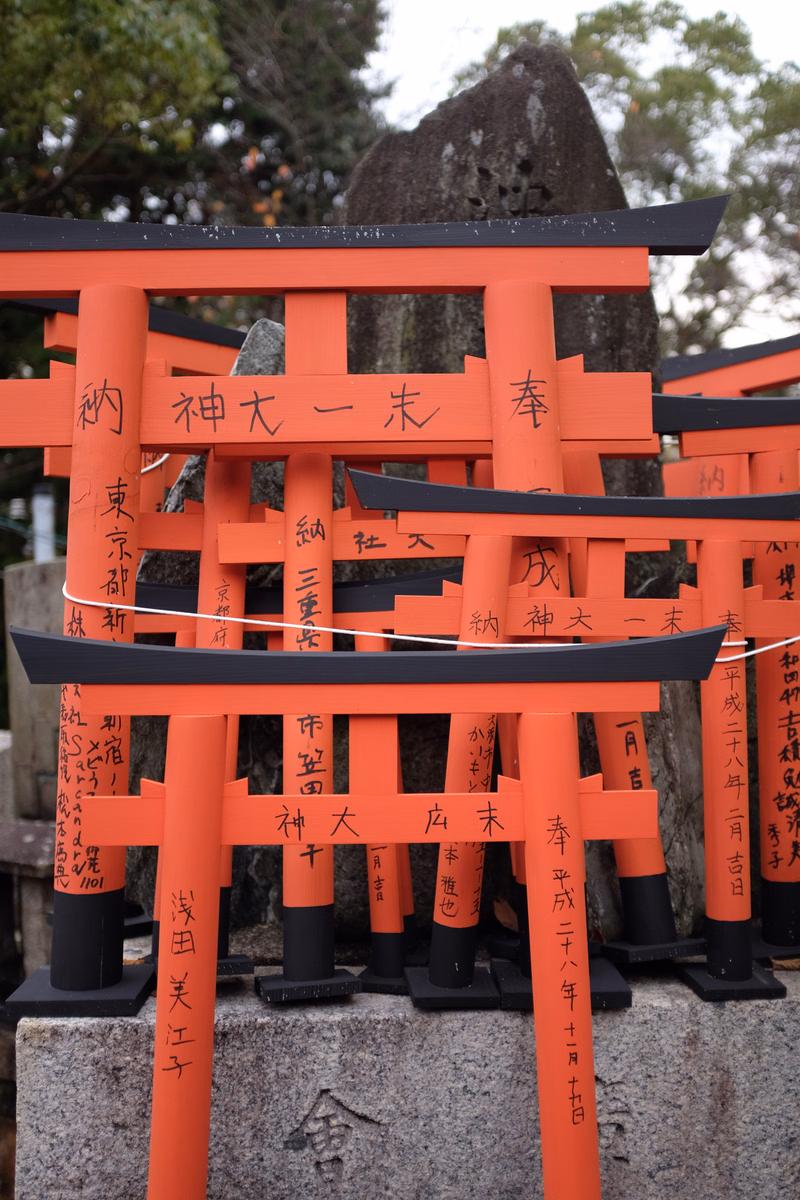 A row of bright orange torii gates, some with Japanese text like "納神大一本春", are lined up in front of a large stone monument.