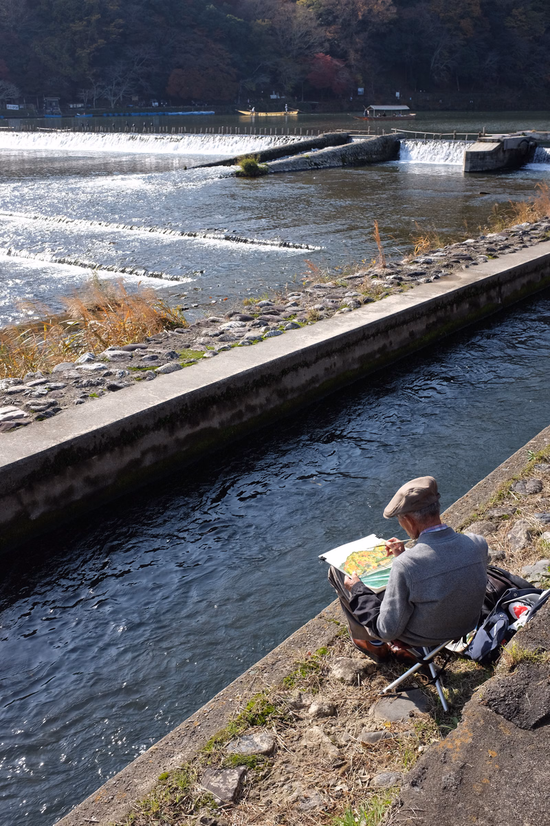A person sits by a concrete canal bank, drawing in a sketchbook while looking out over the flowing water.