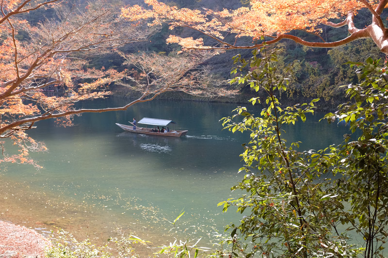 A small boat travels across a calm lake surrounded by trees showing autumn foliage.