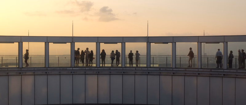 A group of people stands on a balcony overlooking a cityscape during a sunset.