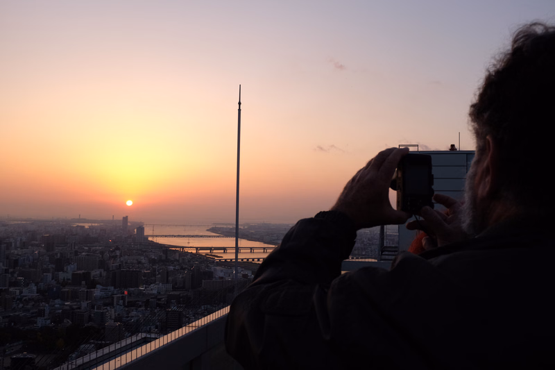A person silhouettes against a vibrant sunset over a sprawling cityscape, holding a camera to take a picture.