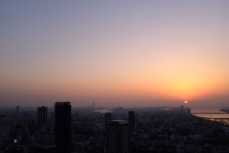 A wide cityscape view at sunset, with the sun setting over the horizon and silhouettes of buildings in the foreground.