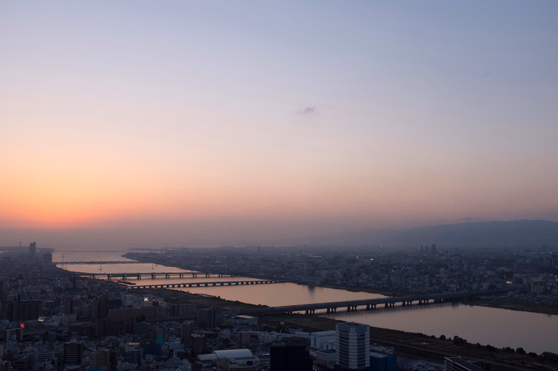 A wide view of a city skyline at sunset, featuring a large river spanned by multiple bridges under a colorful sky.