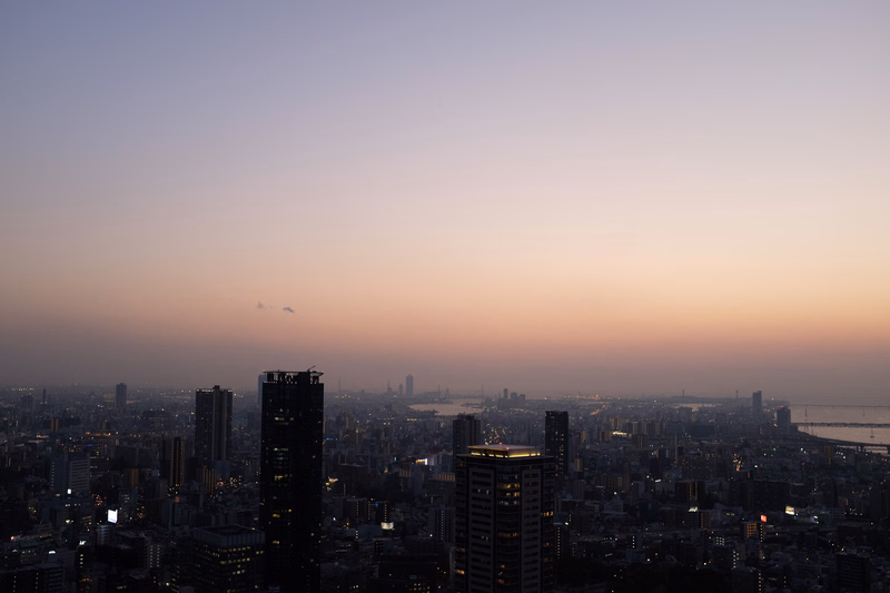 A wide cityscape view at sunset, showing numerous high-rise buildings against a pale orange and purple sky.