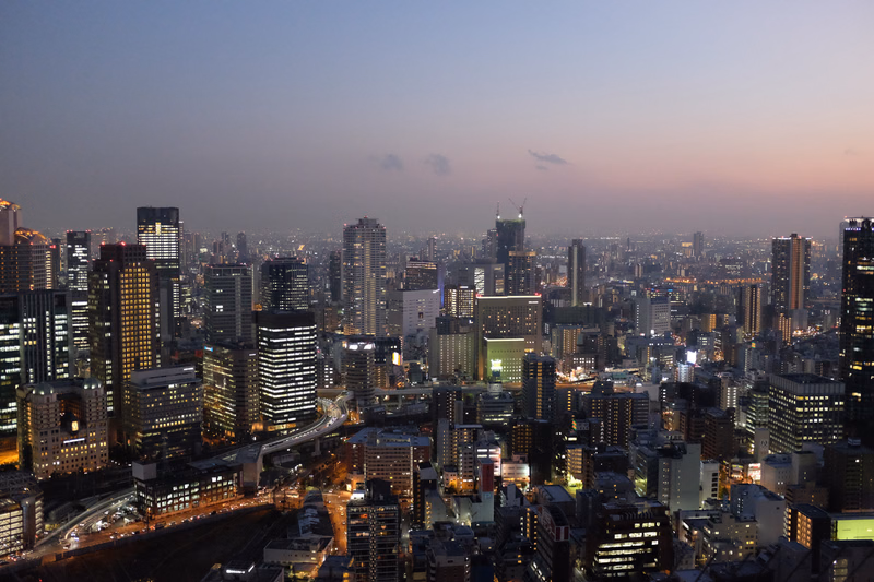 A wide panoramic view of a sprawling city skyline during the twilight hours, with numerous high-rise buildings visible against a pale sky.
