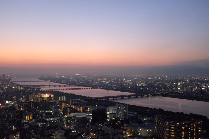 A wide panoramic view of a city skyline at sunset, featuring a large river with bridges crossing the water.