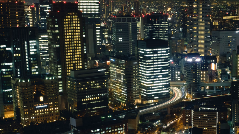 A dense nighttime cityscape view showing numerous illuminated high-rise buildings and busy traffic on elevated roadways.