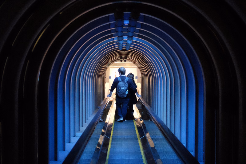 Two people walk down a long, arched, blue-lit tunnel walkway.
