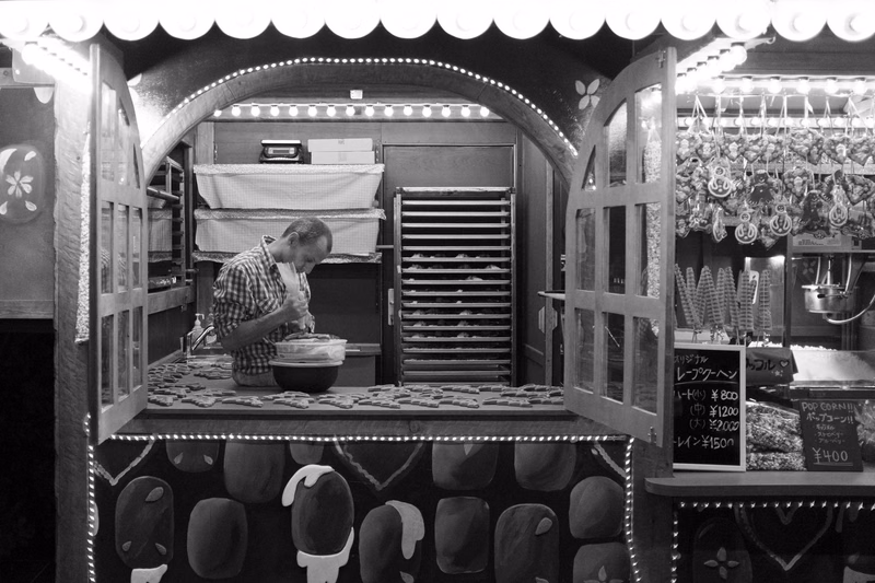 A black and white photo of a beach hut shop with a counter where a man is preparing food, and a display window showing items.