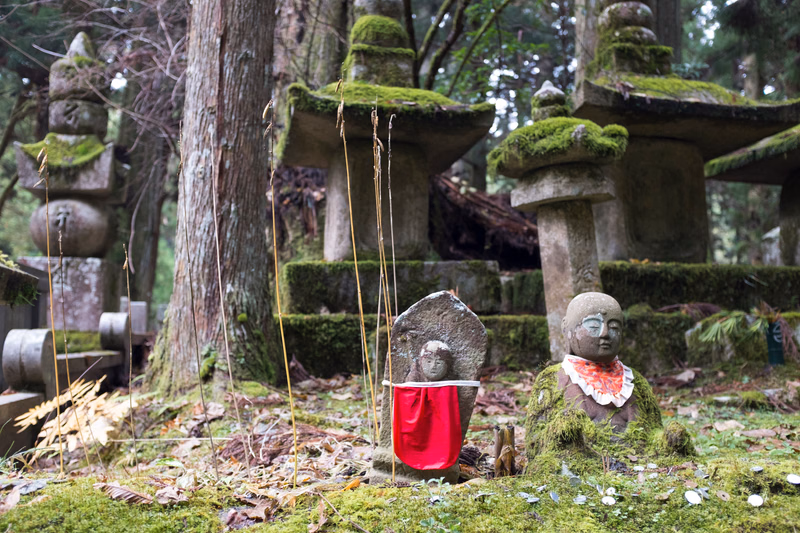 Two small stone figures, one with a red cloth, sit among moss-covered stone structures in a wooded garden setting.