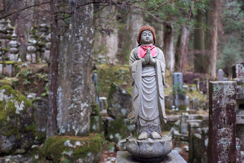 A stone statue of a Buddha figure with pink accents stands in a mossy, wooded garden area surrounded by gravestones.