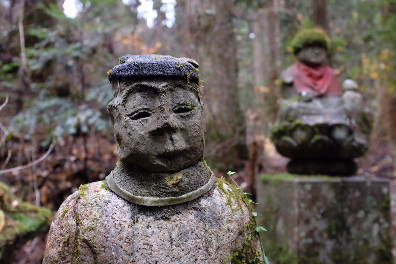A weathered stone statue with moss on its head is in the foreground, with another statue visible in the background amidst trees.