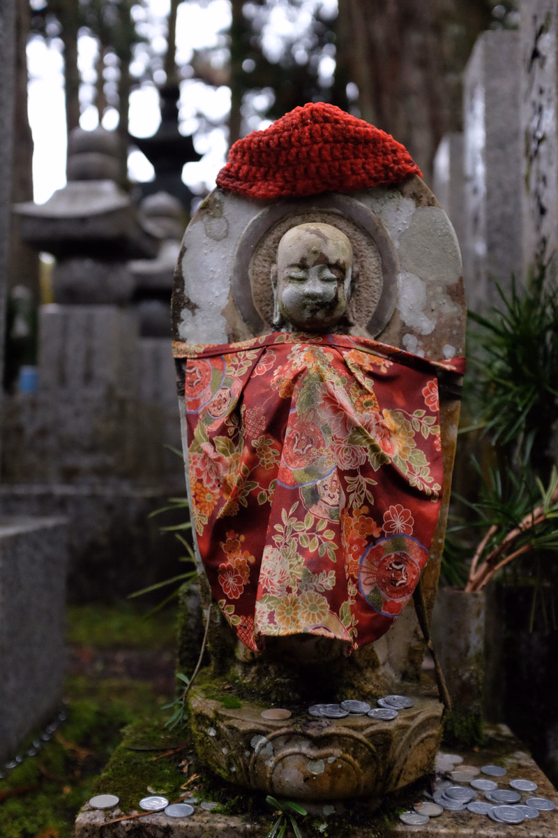 A weathered stone statue of a religious figure wearing a red hat and a patterned red cloth stands in a cemetery.