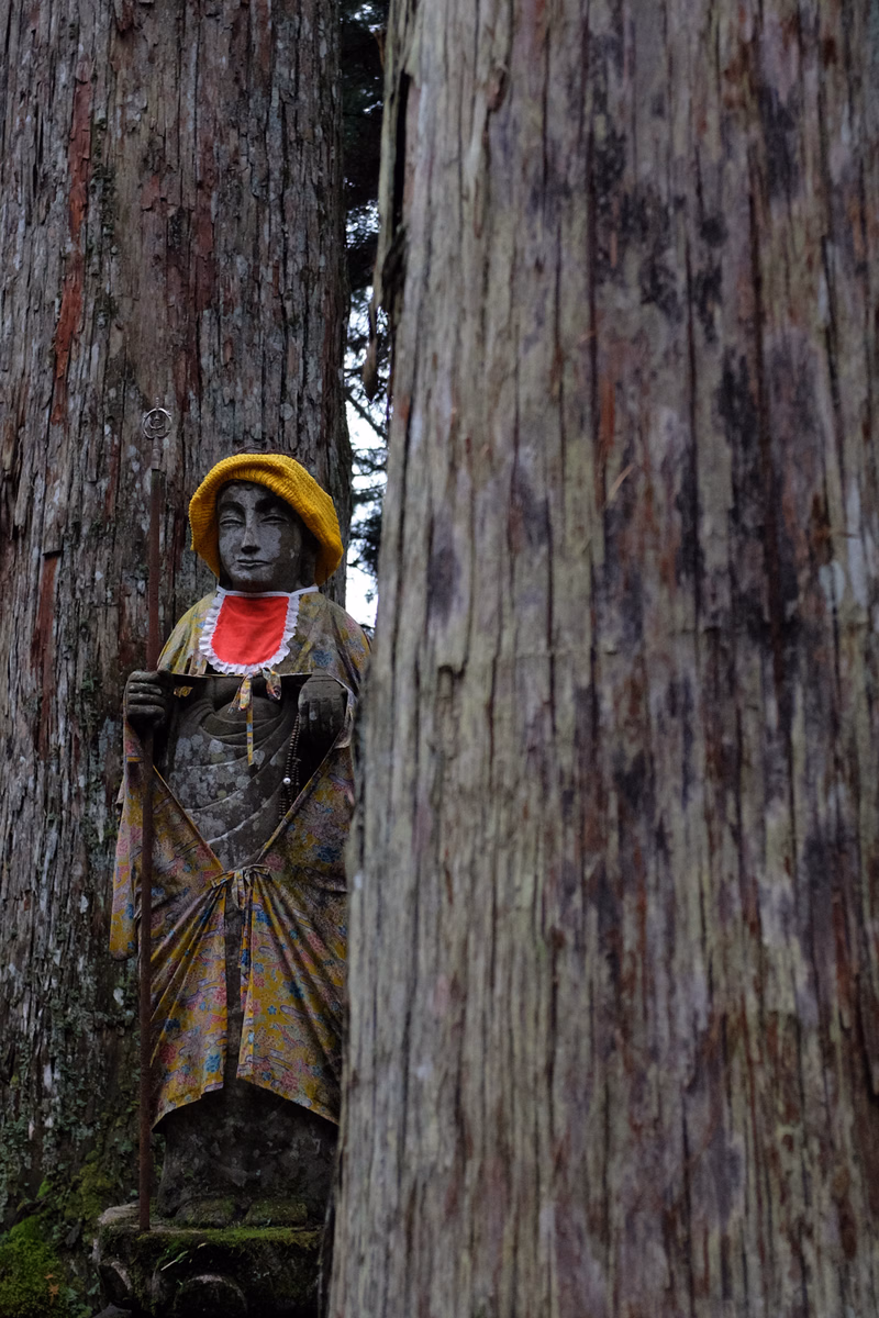 A colorful statue of a figure wearing a yellow hat stands between two large, textured tree trunks outdoors.