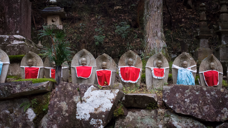 A serene and contemplative scene of stone statues in a forested area, with red cloth draped over them, and a traditional Japanese lantern in the background.
