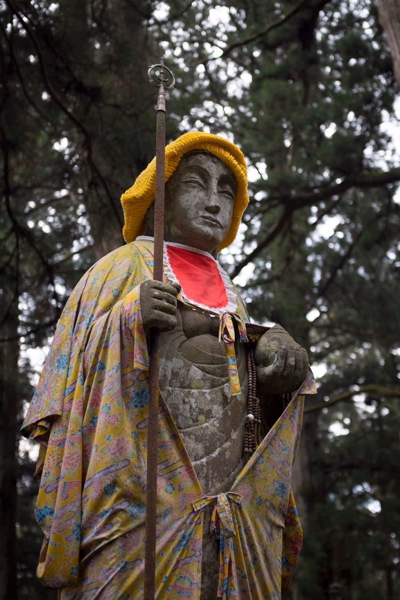 A colorful statue of a figure wearing a yellow hat stands outdoors among tall trees, holding a staff.