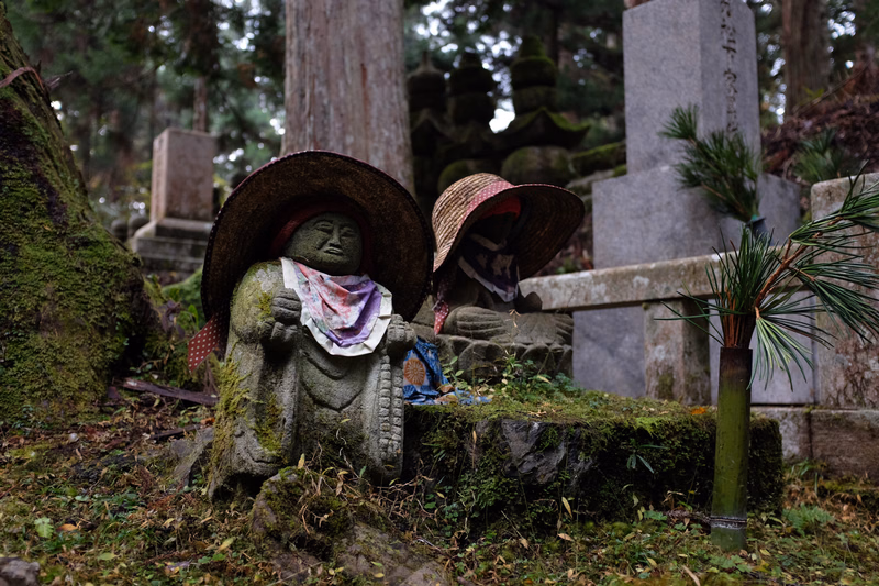 Two moss-covered stone guardian statues wearing wide-brimmed hats and colorful cloths are situated in a damp, overgrown graveyard.