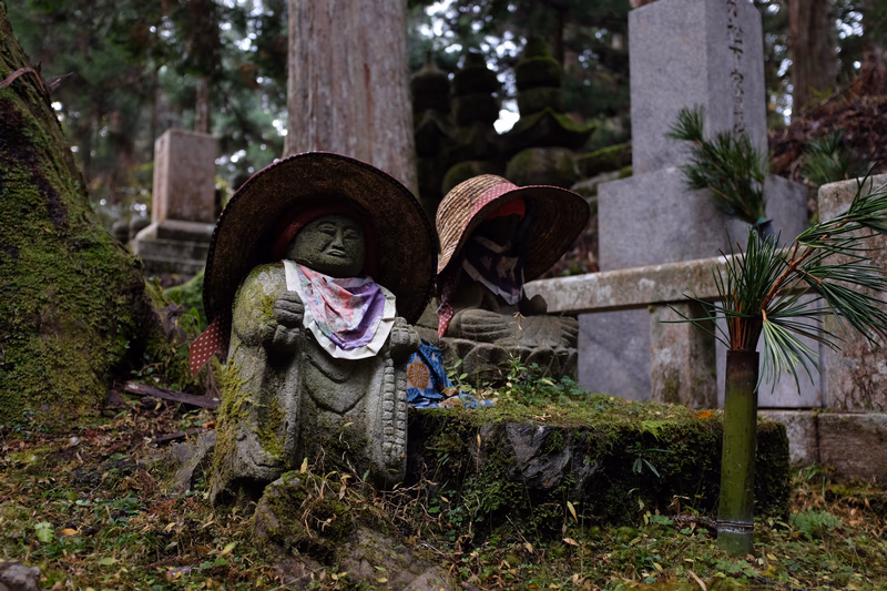 Two stone statues wearing straw hats are placed on a moss-covered stone in a serene cemetery.
