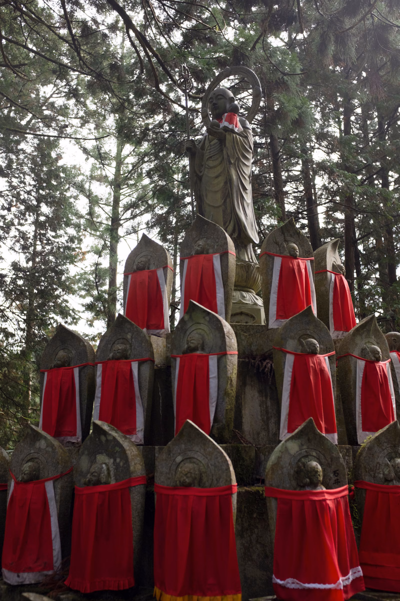A large statue of a religious figure stands among a row of smaller stone monuments and crosses in a wooded area.