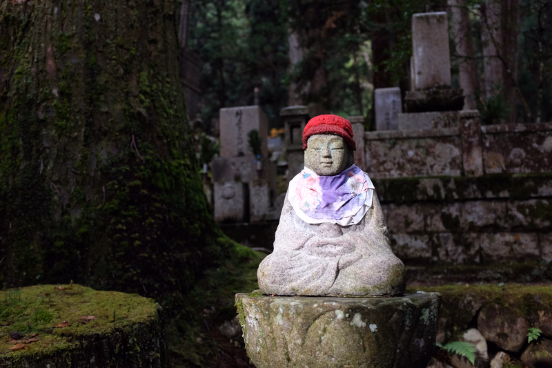 A stone statue wearing a red hat and purple cloth sits in a mossy, shaded garden area with old stone monuments in the background.