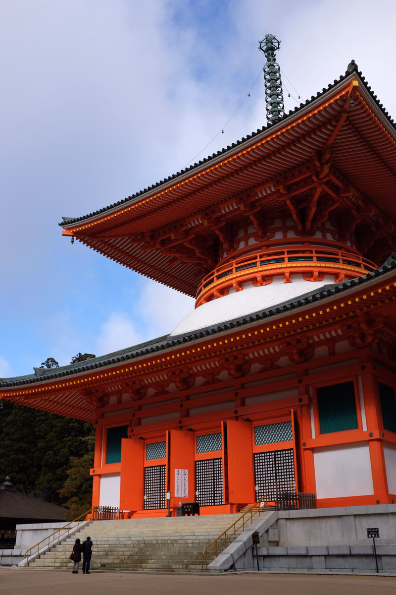 A large, vibrant red Japanese temple building with tiered roofs is shown under a partly cloudy sky.