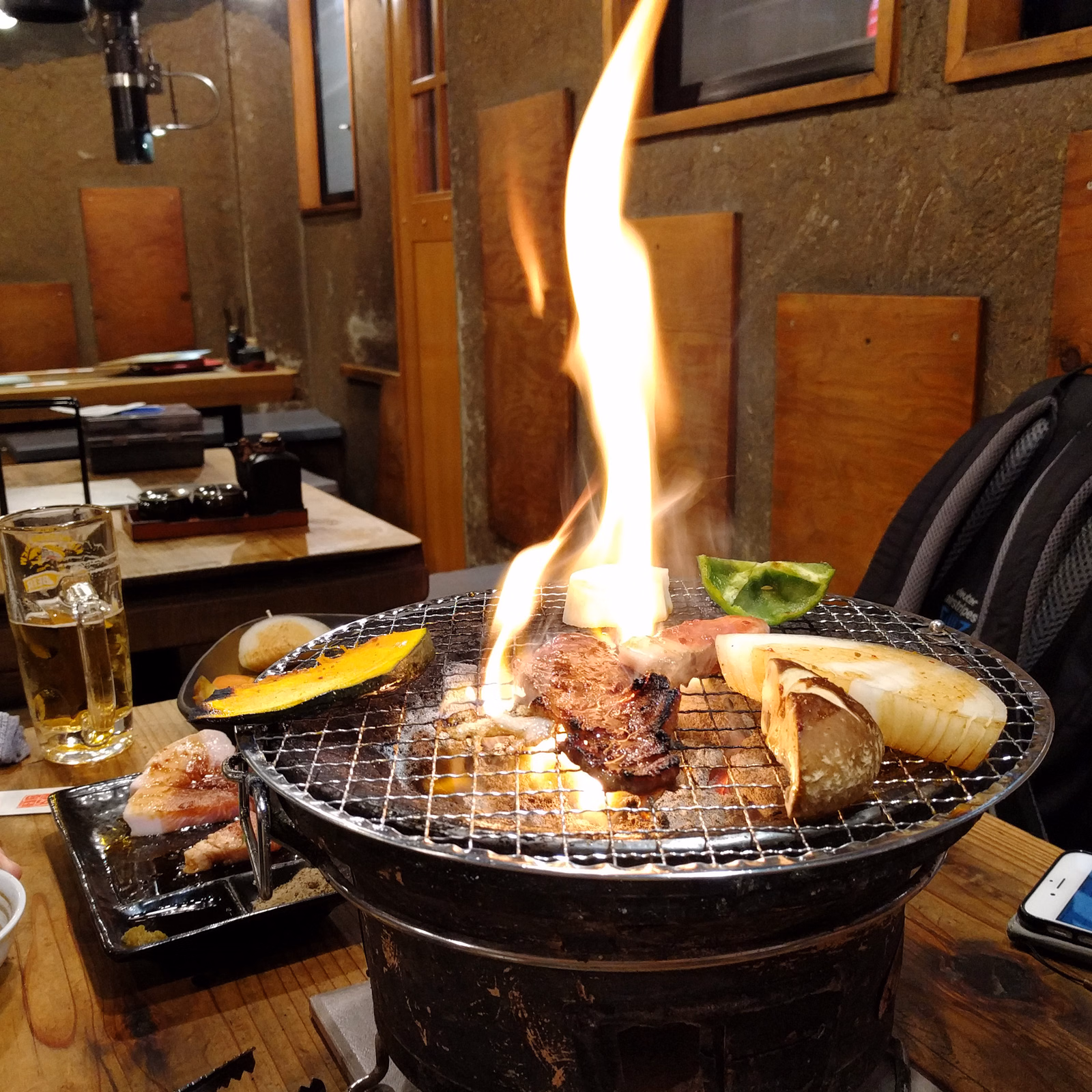 A large metal grill is actively cooking meat and vegetables over a bright, tall flame in an indoor setting.