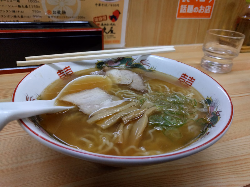 A bowl of ramen noodle soup with slices of pork and green onions, served on a wooden table near a sign that reads "話題のお店".