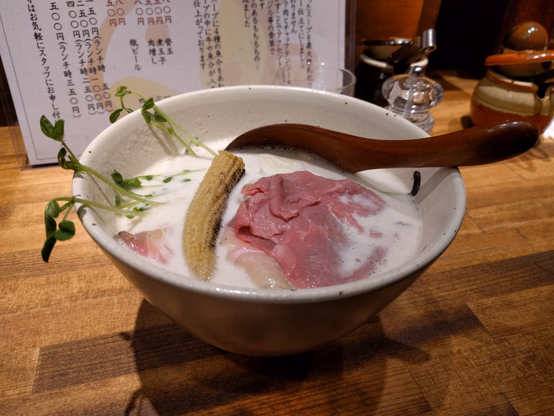 A bowl of soup containing slices of pink meat, garnished with green sprouts, served with a wooden spoon.