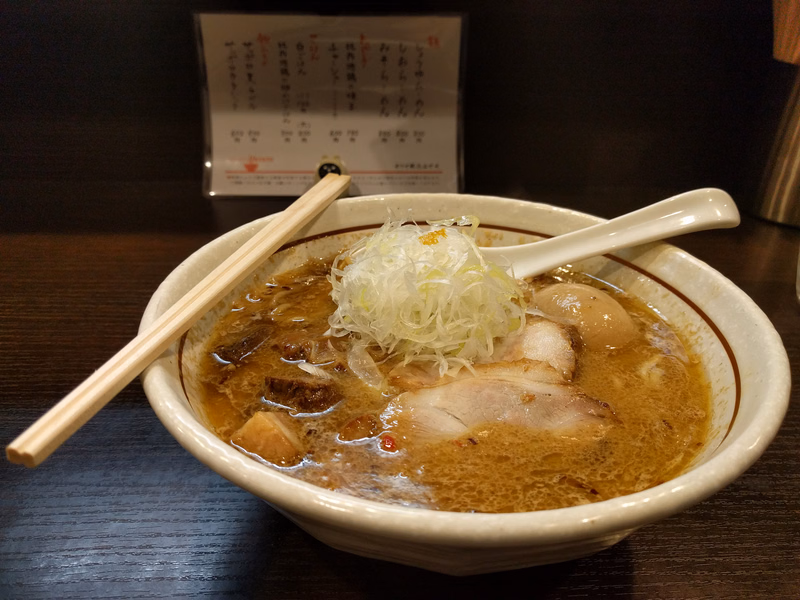 A bowl of tonkotsu ramen with pork slices and white onion garnish sits on a dark wooden table, next to a menu.