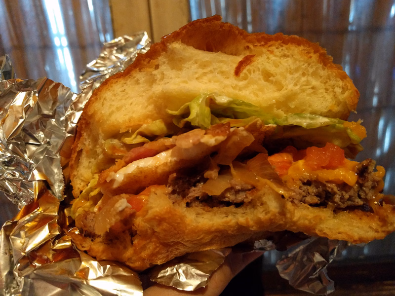 A large, hearty burger with meat, cheese, lettuce, and tomato is served on a toasted bun, wrapped in aluminum foil.