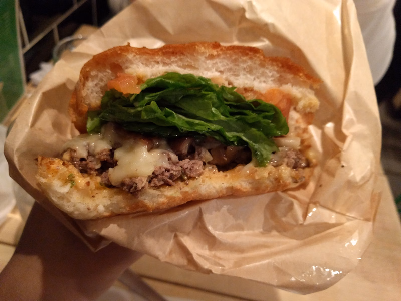 A grilled sandwich with meat, melted cheese, and fresh green lettuce is served in a brown paper wrapper.