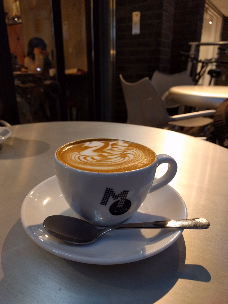 A white cup of latte art sits on a saucer on a light-colored table in a cafe.