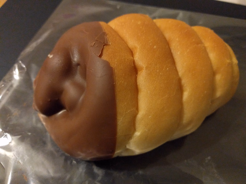 A golden brown sweet roll with a thick layer of smooth chocolate frosting is resting on a clear plastic surface.