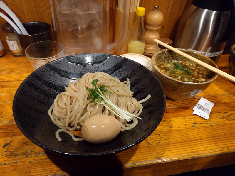 A black bowl of udon noodles with a boiled egg sits on a wooden table next to a bowl of miso soup.