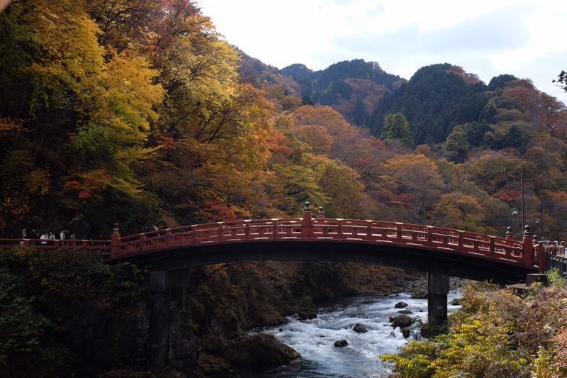 A red arched bridge spans a rocky river surrounded by vibrant autumn foliage and forested mountains under a cloudy sky.
